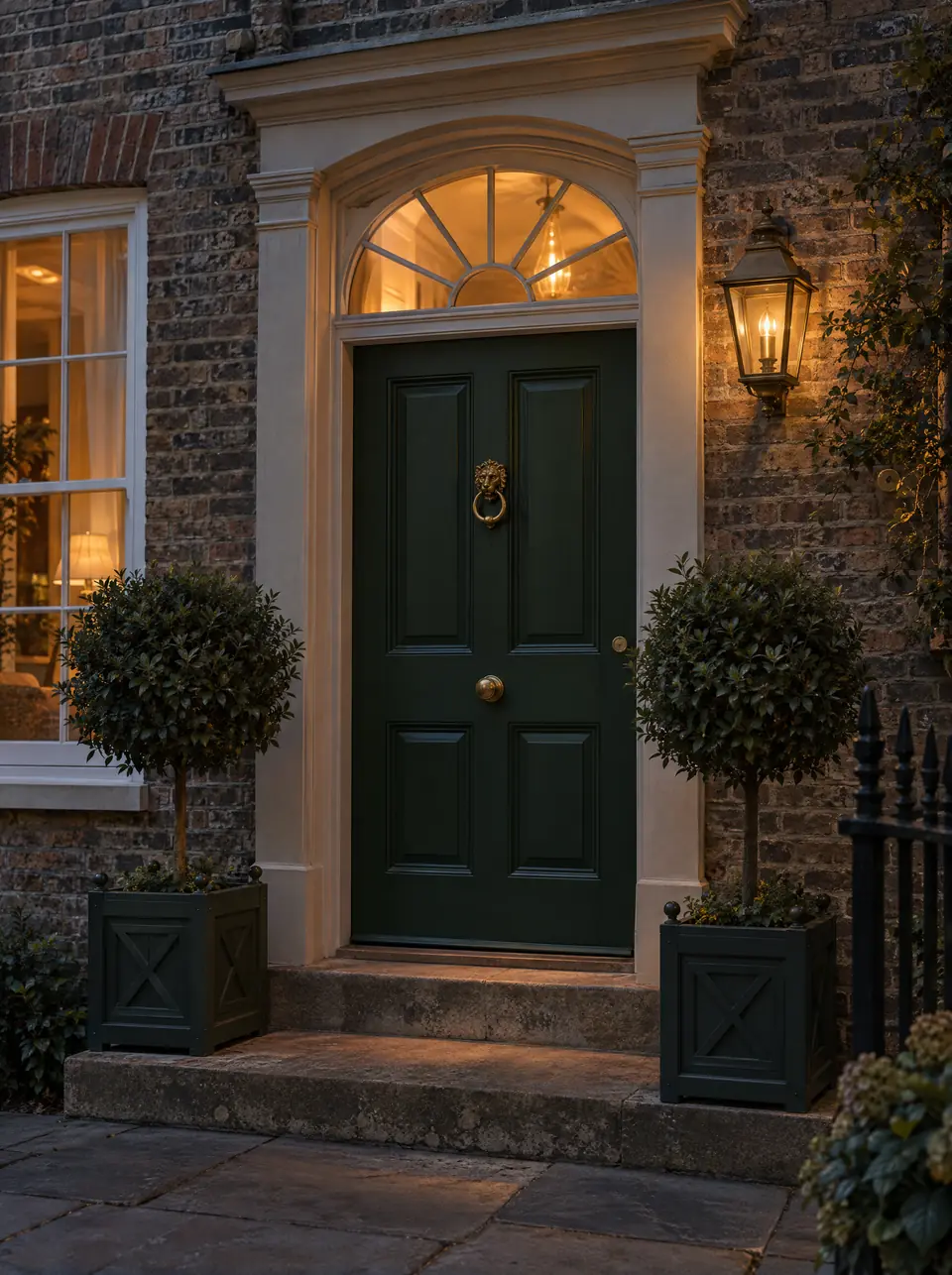 A dark green panelled front door with brass knocker, lit by a lantern at dusk between two clipped bay trees.